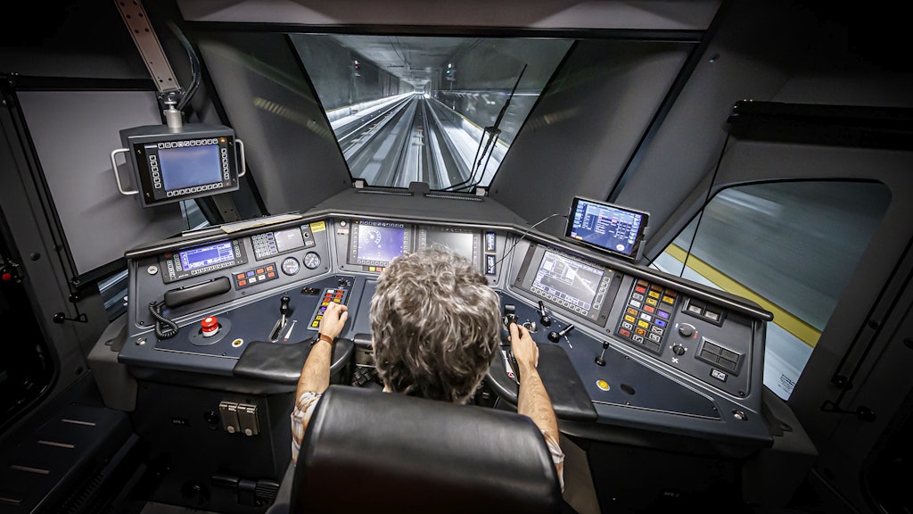 Sguardo nella cabina di guida di una moderna locomotiva, mentre attraversa una galleria. Il “tavolo da lavoro” del macchinista è costituito da più schermi e da spie e tasti colorati. Si tratta del veicolo motore di un treno Flirt del Leman Express.