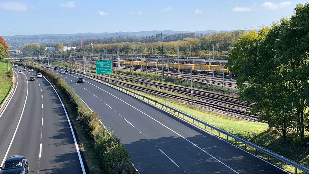 Un tratto autostradale a quattro corsie costeggiato da binari. Su un binario transita un treno della Posta, con il Giura sullo sfondo. 