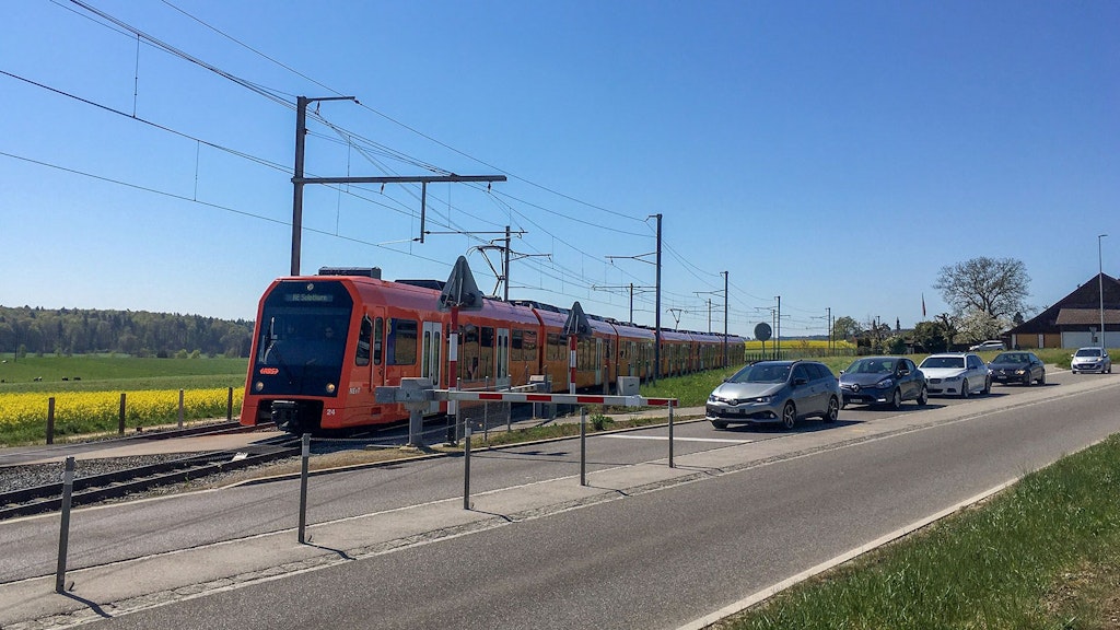 Un treno arancione della RBS e una colonna di auto ferme davanti a una barriera.