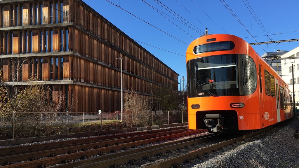 Un train orange du RBS passe devant le bâtiment de l’OFT dont la façade est en bois.