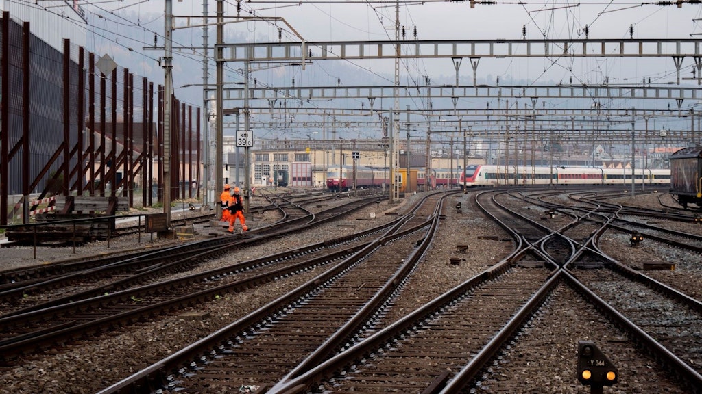 Mehrere Bahngleise mit Oberleitungen, zwei Bahnarbeiter in orangefarbener Kleidung und ein moderner Zug im Hintergrund.