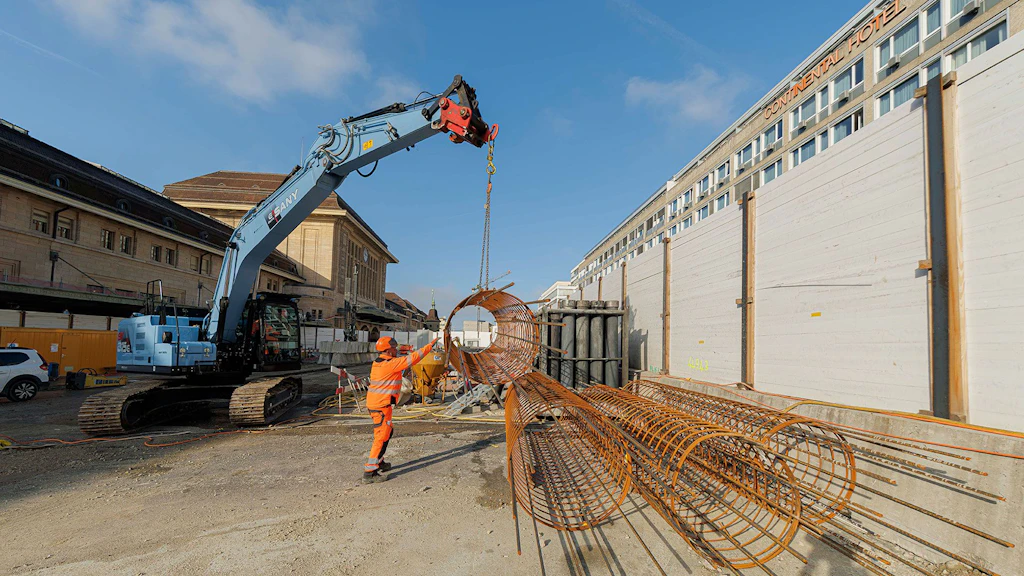 Elektrischer Bagger hebt auf einer Baustelle eine Stahlarmierung, gesteuert von einer Person in Sicherheitskleidung. 
