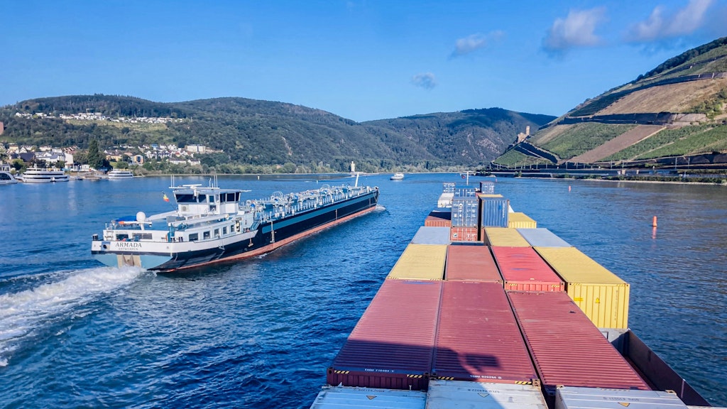 Unter einem blauen Himmel schwimmt ein Frachtschiff auf dem Rhein, das von einem Passagierschiff überholt wird. Rechts davon ein Weinberg.