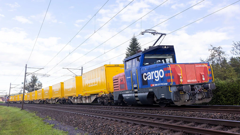 Ein Güterzug der SBB Cargo mit gelben Güterwagen der Post fährt durch eine ländliche Umgebung.