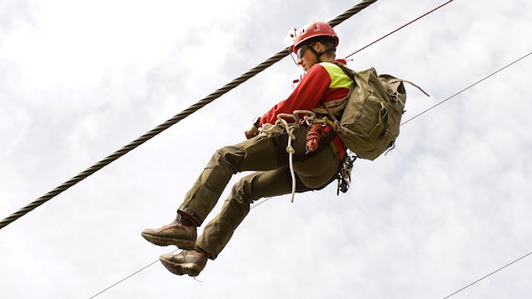 Ein Kontrolleur schwebt an einem Seilbahnseil befestigt in der Luft.