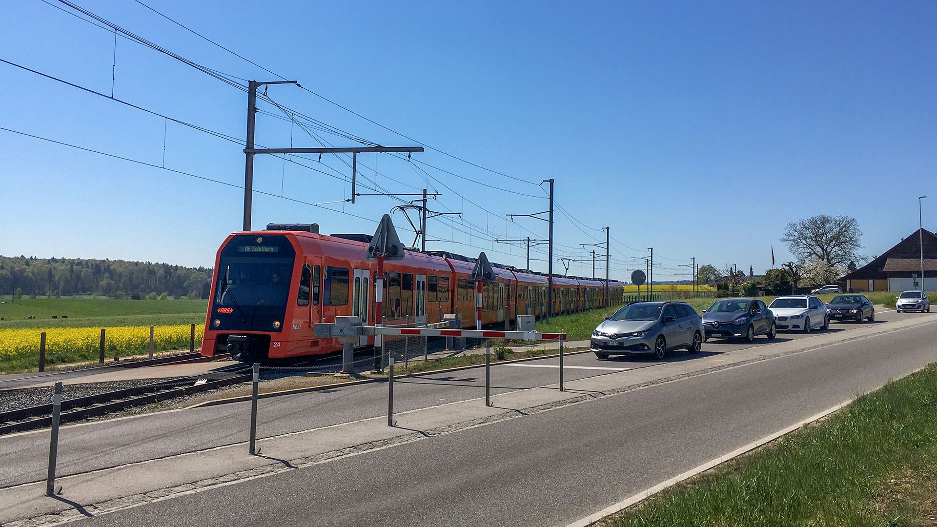 Ein oranger RBS-Zug fährt an vor einer Barriere wartenden Autos vorbei.