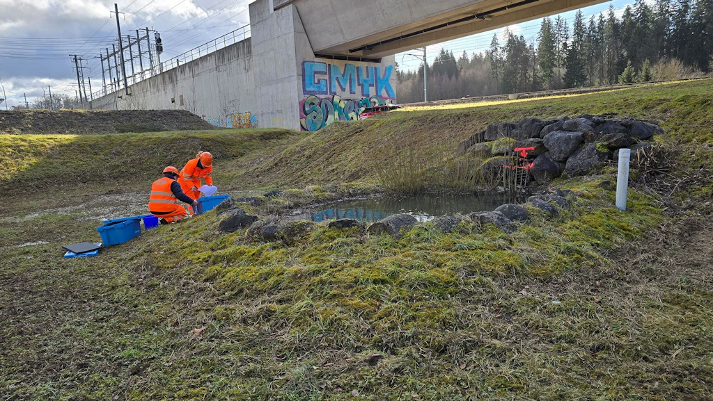 Zwei Personen in Warnwesten an einem Teich in der Nähe von Gleisanlagen.