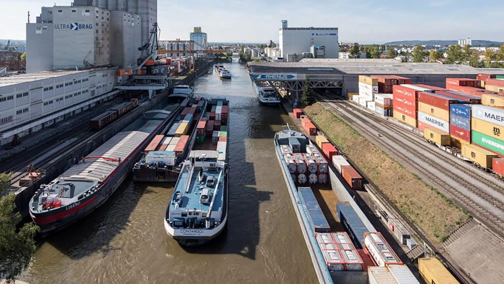 Das Bild zeigt einen Hafenkanal in Kleinhüningen mit mehreren Frachtschiffen, die mit Containern beladen sind. Links und rechts des Kanals sind Eisenbahngleise sichtbar. Im Hintergrund am Ufer des Kanals ein hohes graues Betongebäude.