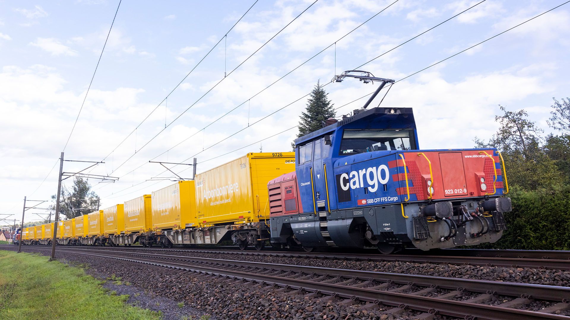 An SBB Cargo freight train made up of yellow Swiss Post freight wagons travels through a rural area.