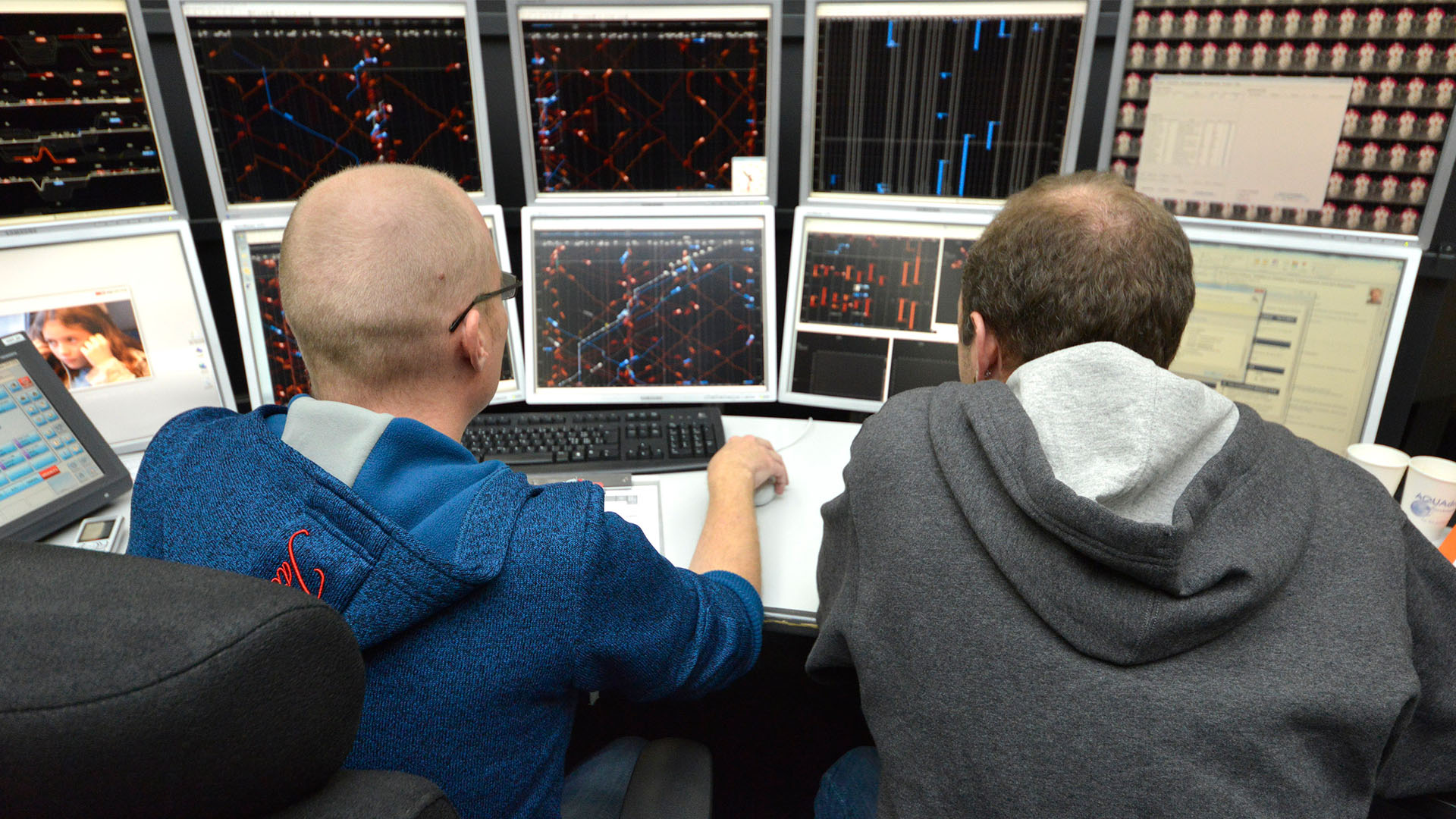 Two men sit in front of ten monitors showing red and blue lines.