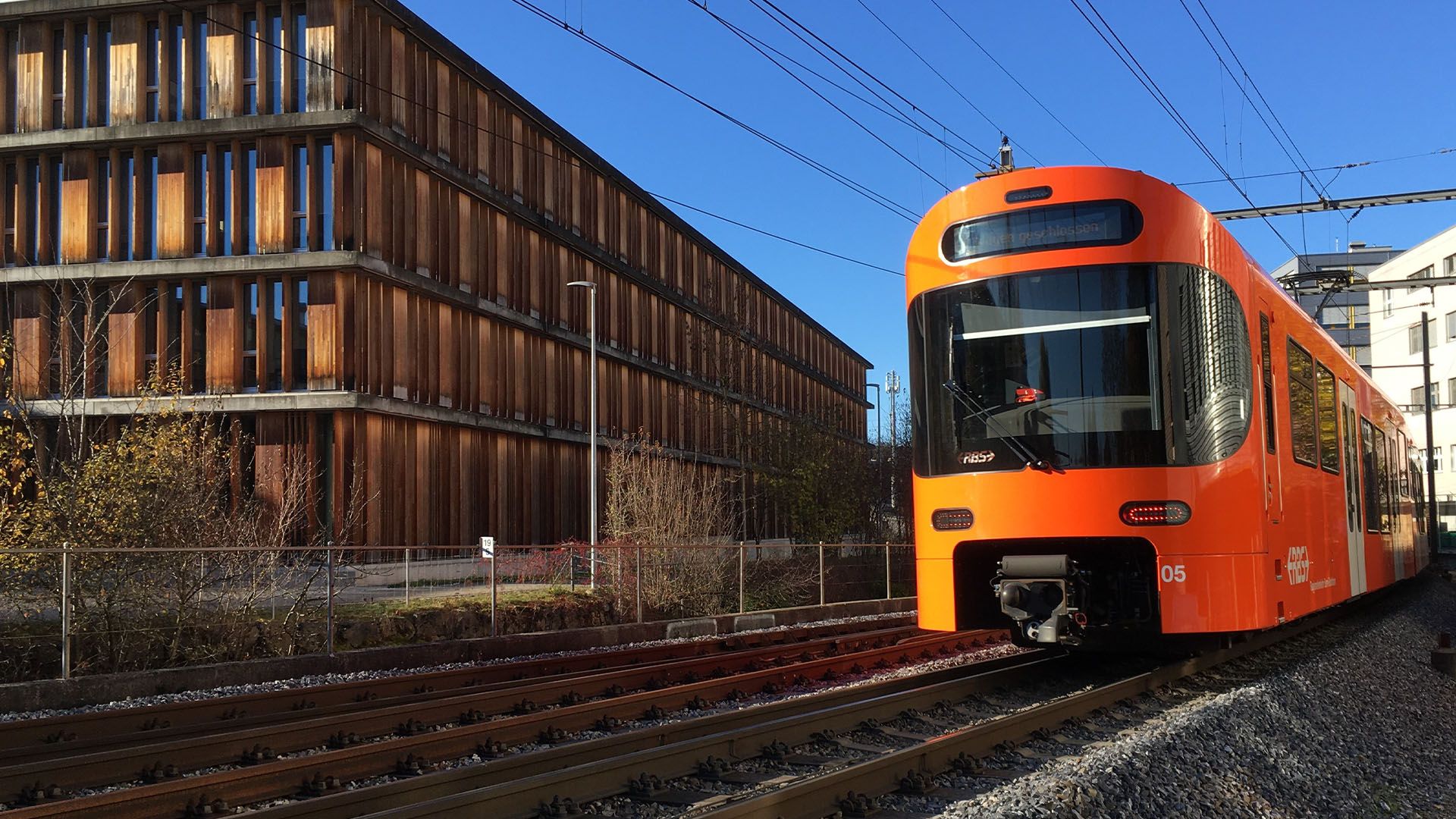 An orange RBS train runs past the timber-clad BAV building.