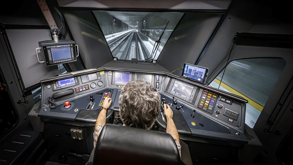 Vue de l’intérieur d’une cabine de conduite d’une locomotive moderne qui traverse un tunnel. Plusieurs écrans, des voyants lumineux et des boutons de plusieurs couleurs délimitent le plan de travail du conducteur de locomotive. Le véhicule moteur fait partie d’une rame Flirt du Léman Express.