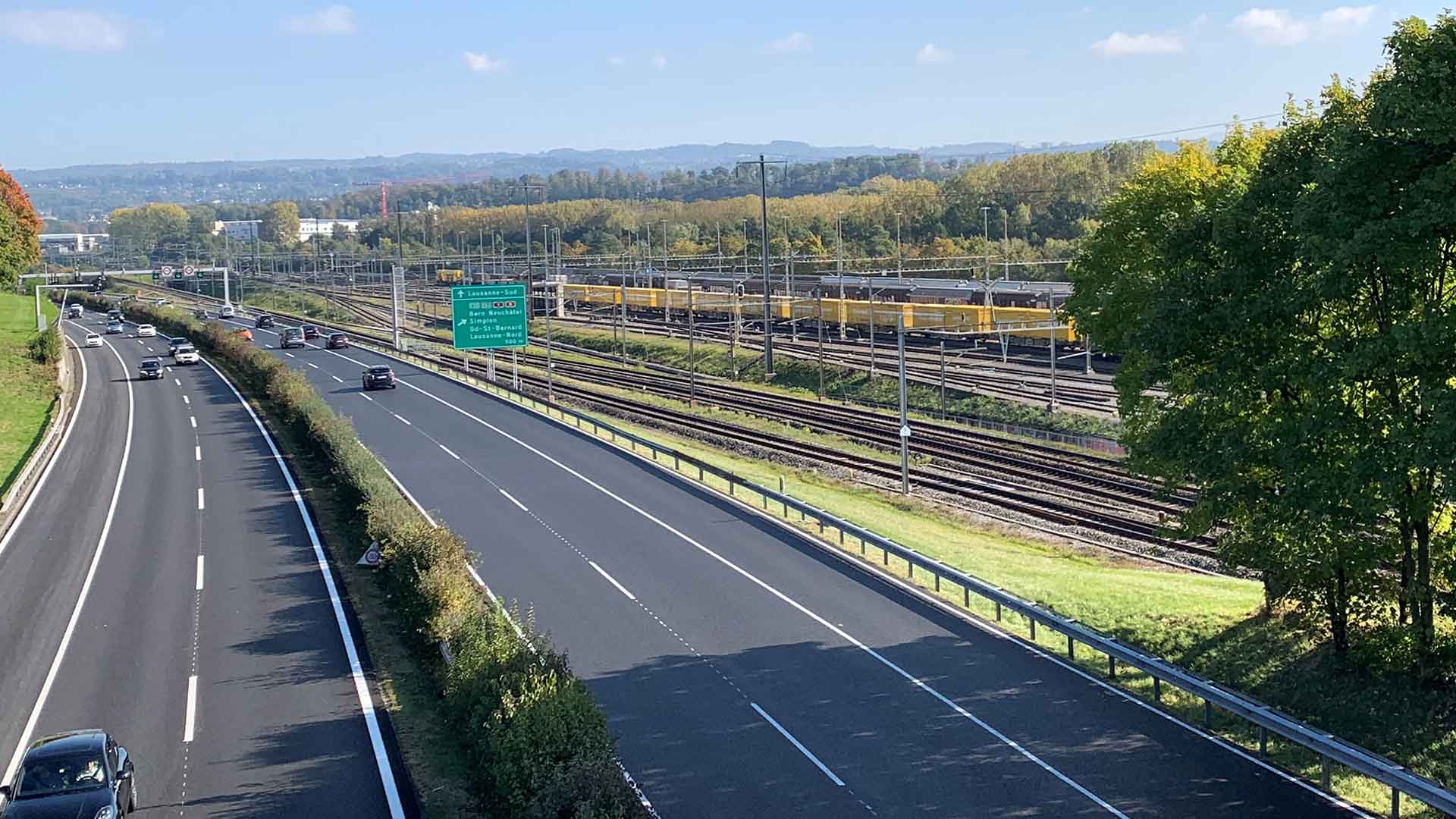Une autoroute à quatre voies et, à droite, de nombreuses voies ferrées. Un train de la Poste circule sur l’une des voies. En arrière-plan, le Jura.