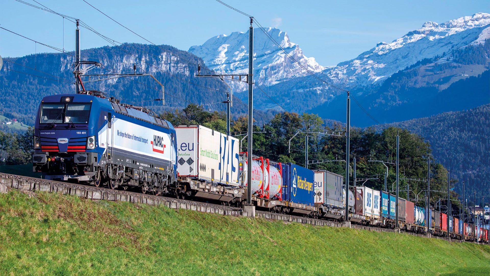 Un treno merci in transito sullo sfondo di un paesaggio alpino.